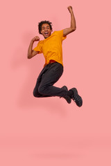 Fototapeta na wymiar Full length photo of cheerful African American man jump raise fists to scream yes celebrate a victory or rejoicing with sales starting, wear orange casual t-shirt isolated over pink background