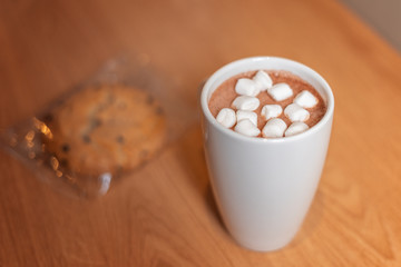 A mug of cocoa and marshmallow on a wooden table next to an oatmeal cookie.