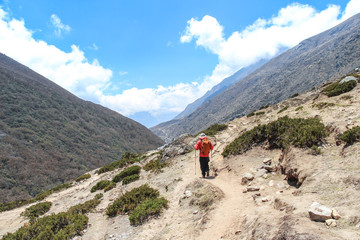 Fototapeta na wymiar Back view. Man in red windbreaker with orange backpack walks on footpath in Himalayas on the way back from Everest base camp. Clouds lies on mountainside. Theme of trekking in Nepal.