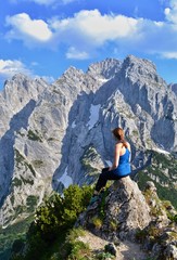 Young girl is sitting on the top of the mountains, enjoying beautiful view on the mountain range Kaisergebirge, Austria. Sunny day, white clouds, blue sky. 