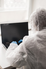 Girl in medical mask and white transparent protective suit sits in quarantine at home and works at computer at the table. Designer, artist, architect at remote work in a pandemic covid.