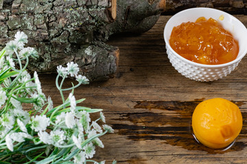 Apricot and peach jam in a small deep dish, sweet orange jam for dessert. Next to a whole fruit, apricots are marinated in syrup. Old wooden background and snags with bark with white flowers