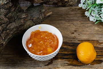 Apricot and peach jam in a small deep dish, sweet orange jam for dessert. Next to a whole fruit, apricots are marinated in syrup. Old wooden background and snags with bark with white flowers