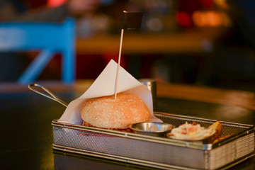Big burger with baked potatoes, salad nad sauce in a tray on dark wooden table over blurred background in a restaurant.