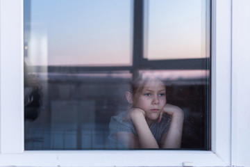 A sad, lonely child sits on the windowsill and looks out the window. Stay at home quarantine coronavirus prevention of the pandemic. The view from the street. Prevention of the epidemic. - obrazy, fototapety, plakaty