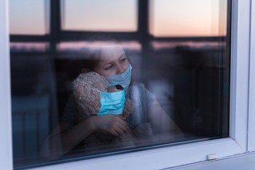 Stay at home quarantine coronavirus pandemic prevention. Sad child and his teddy bear both in protective medical masks looks out window. View from street. Prevention epidemic.