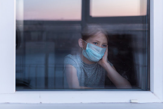 Stay At Home Quarantine Coronavirus Pandemic Prevention. Sad Child In Protective Medical Mask Looks Out Window. View From Street. Prevention Epidemic.