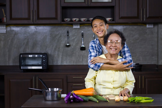 Happy Young Aisan Man Hugging His Grandmother While Prepare Various Of Vegetable To Cooking At Home Kitchen. Healthy Lifestyle And Domestic Life Of Asian People Concept.