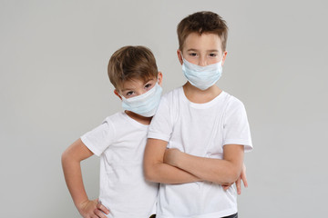 Two cute little boys in white clothes and protective medical masks stand in a hug isolated on a gray background.