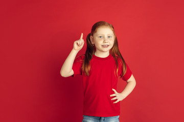 Pointing up. Caucasian little girl portrait isolated on red studio background. Cute redhair model in red shirt. Concept of human emotions, facial expression, sales, ad, childhood. Copyspace.