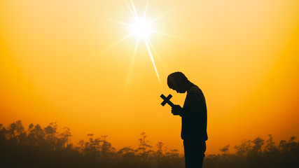 Young man praying with holding christian cross in morning light, christian silhouette concept.