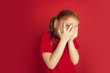 Scared hiding face. Caucasian little girl portrait isolated on red studio background. Cute redhair model in red shirt. Concept of human emotions, facial expression, sales, ad, childhood. Copyspace.