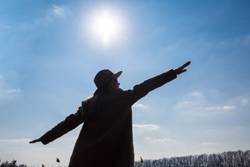 Silhouette of a girl in a hat and coat on a background of the sky with clouds and the sun.