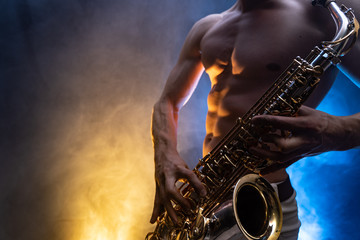 Muscular man with naked torso playing on saxophone with smoked colorful background 