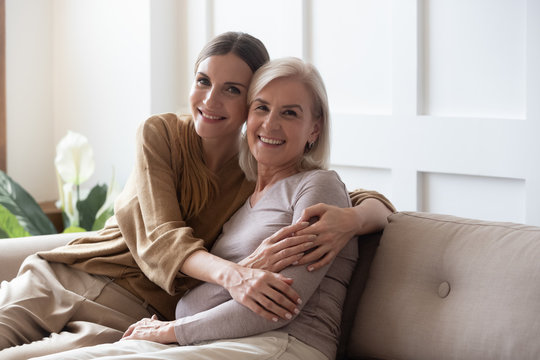 Portrait happy adult daughter and older mother hugging and holding hands, sitting on couch at home, young woman and mature mum or grandmother looking at camera, two generations family photo