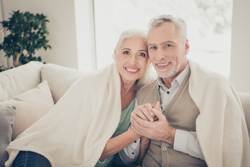 Perfect weekend in new house. Close up photo of charming grandparents holding hands sharing beaming smile sitting on soft divan