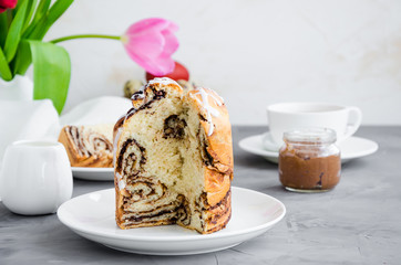 Easter background. Easter cake - Cruffin, Kraffin or Kulich with chocolate nut cream and sugar glaze on top on a white plate on a concrete background. Horizontal orientation. Copy space.