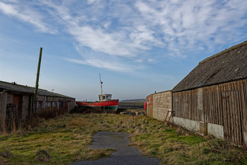 An old traditional wooden Fishing boat up in blocks and out of the water between old wooden and derelict sheds and garages.