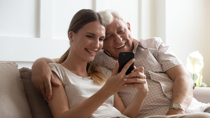 Smiling older father with adult daughter using phone together, having fun, watching video in social...