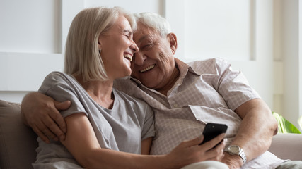 Happy older wife and husband cuddling close up, using phone together, laughing at funny video in...