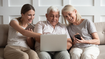 Happy older parents with adult daughter using electronic devices, phones, looking at laptop screen,...