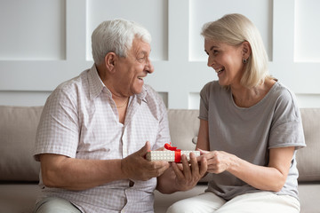 Smiling older wife congratulating husband with birthday, giving box with red bow, happy adult middle-aged daughter presenting gift to mature father, family celebrating fathers day, two generations