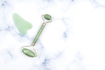 Green face roller and gua sha massager made from natural jade nephritis stone over marble background.