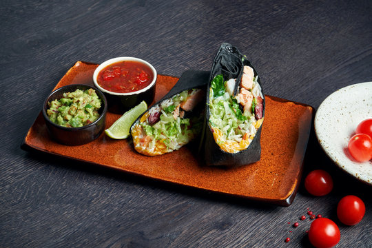 Close Up View On Burrito With Salmon, Lettuce, Rice, Tomatoes, Corn And Bell Pepper In Black Pita On A Brown Plate With Tomato Salsa And Guacamole. Vegetarian Shawarma Roll