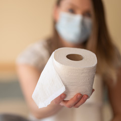 The masked girl is holding toilet paper against the background of the room. The paper is in focus. Panic and mass buying toilet paper. Coronavirus