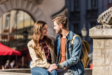 Fototapeta premium Happy girlfriend and boyfriend sitting together, looking at each other and smiling in city