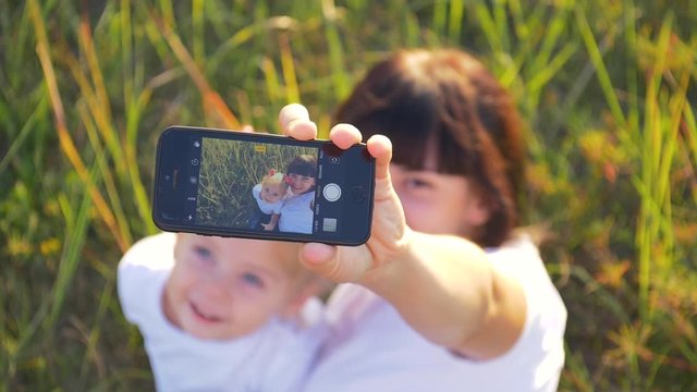 Family mom and daughter take selfie on smartphone sitting on green grass in summer day.