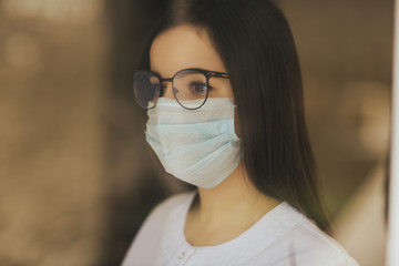 Home quarantine. Woman is sitting at window in a medical mask and glasses , looking out, wants to get out. Protection against coronavirus infection, pandemics. COVID-19. 