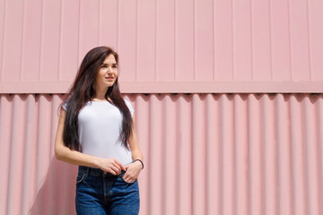 Cute young woman with long straight dark hair in white t-shirt and blue jeans on pink background. Female standing tucked hands in her pockets against colored iron wall. Copyspace. Fashion summer photo