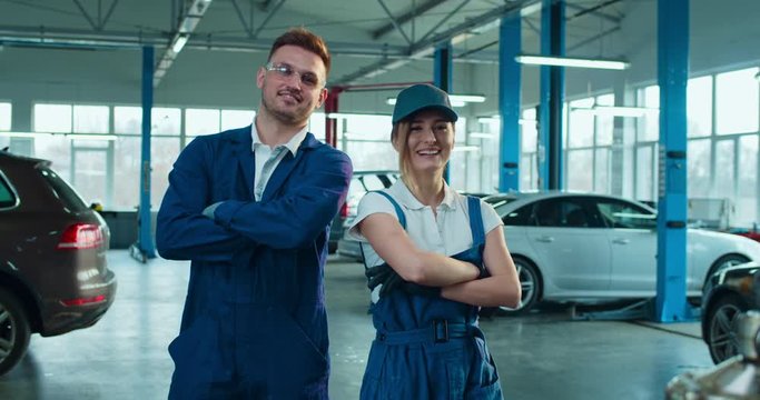 Couple of male and female auto mechanics in uniforms giving five to each other and laughing joyfully. Portrait of pretty woman and handsome man smiling to camera in big garage.