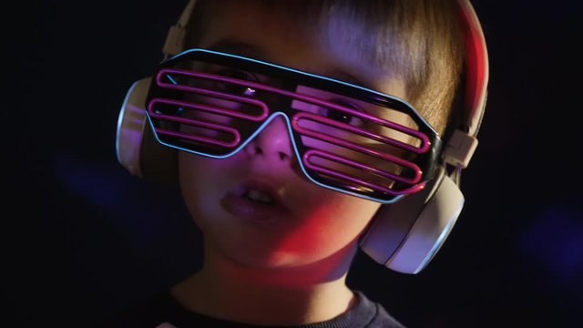 Portrait of a little boy 5 years old in wireless headphones and stylish neon glasses listens to music, sings.