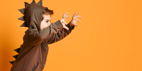 Kid boy in hoodie with dinosaurus spikes at his back and cowl playing t-rex holding hands up demonstrating claws