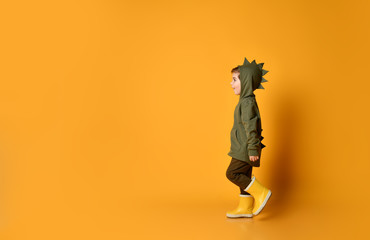 Nice kid boy in hoodie with dino spikes at back and cowl and in yellow rubber boots is walking...