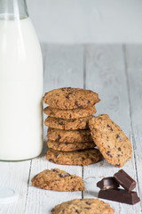 chocolate cookies stacked next to a milk bottle on top of a white wooden board, with pieces of chocolate and space for text