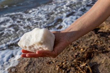 Close-up photo of beautiful and gentle female hand with big piece of sea salt from Dead Sea, Kingdom of Jordan, Middle East. Fine winter sunny day with blurred sea wave and sand in the background