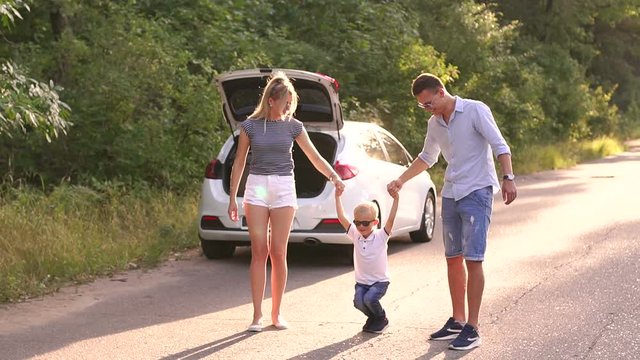 Young parents raise their young son holding hands on an empty road in the woods. A little boy jumps with his parents on the road near the car with trunk open. Family having fun during the trip by car.
