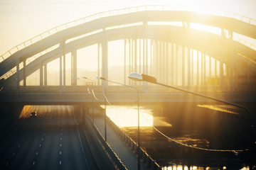 A blurry image of a curved metal bridge over a river, through which the sun's rays pass.