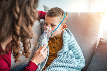 Woman with son doing inhalation with nebulizer at home. Causian little boy making inhalation with nebulizer. Child having respiratory illness helped by mother with inhaler