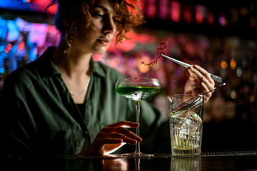 young woman bartender holds glass with cocktail and gently decorates it