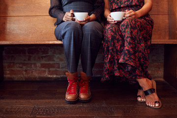 Close Up On Feet Of Female Friends Sitting On Bench In Coffee Shop Together
