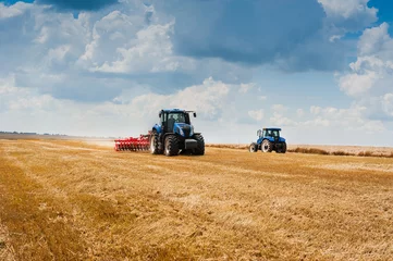 Cercles muraux Tracteur deux tracteurs bleus tirent des herses préparant des terres arables, des champs et de beaux nuages
