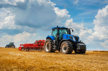 blauwe nieuwe tractor met rode eg in het veld tegen een bewolkte hemel, landbouwmachines werken