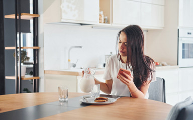 Pouring fresh milk into the glass. Young beautiful brunette in casual clothes indoors in kitchen at daytime