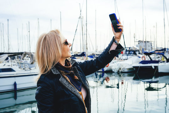 Portrait of an adult lady in sunglasses standing in a marina on yachts background and makes selfie on smartphone.