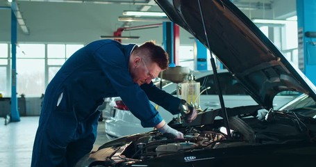Caucasian young male auto mechanic standing at open car and repairing motor with light in big garage. Guy in uniform and goggles working inside automobile repairment salon. Cars service concept.