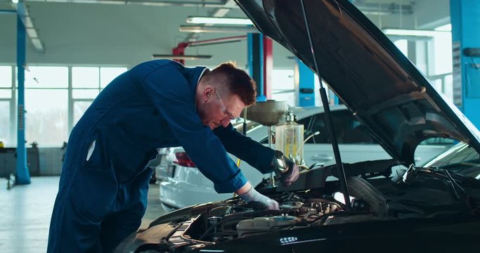 Caucasian young male auto mechanic standing at open car and repairing motor with light in big garage. Guy in uniform and goggles working inside automobile repairment salon. Cars service concept.